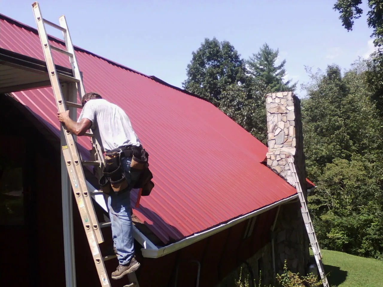 Roofer on a ladder beside a red metal roof and stone chimney — Soffit & Fascia Repair by Roofing Brothers in Davy